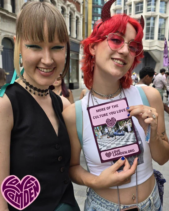 Two people smile to camera. One has bright red hair to shoulder, wearing devil horn headband and loveheart sunglasses. The other person has fringe and bright winged eyeliner with gem stones under eyes. They are holding up a Love Camden flyer.