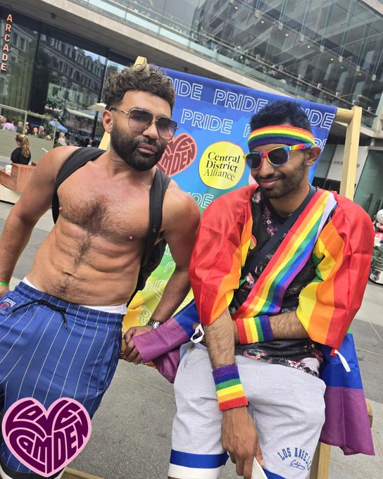Two people wearing sunglasses look to camera, sitting on a PRIDE Love Camden branded deckchair. One of them is wearing rainbow headband and flag around them.