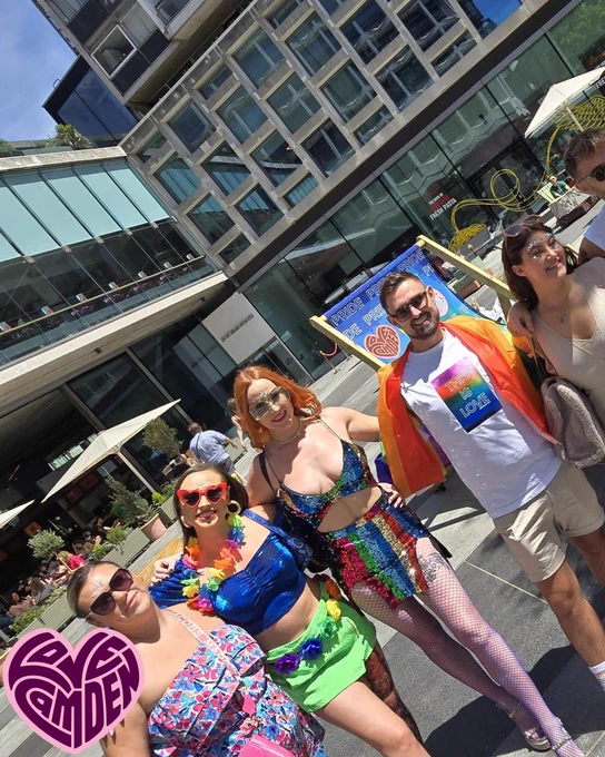 group of 5 people wearing brightly coloured clothes, smiling to camera. One is wearing a sequin rainbow skirt and cropped top, one is wearing a blue crop top and green skirt with floral necklace and another wears a rainbow shirt over white shirt and beige shorts.