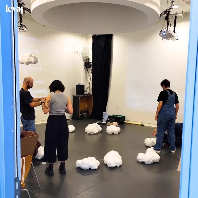 A group of four people are in a room. The camera looks into the room through a blue door frame. The walls of the room are white and the floor is grey. On the floor are clouds made from cotton wool. It looks like the people are setting up an exhibition and the clouds are going to be part of that.