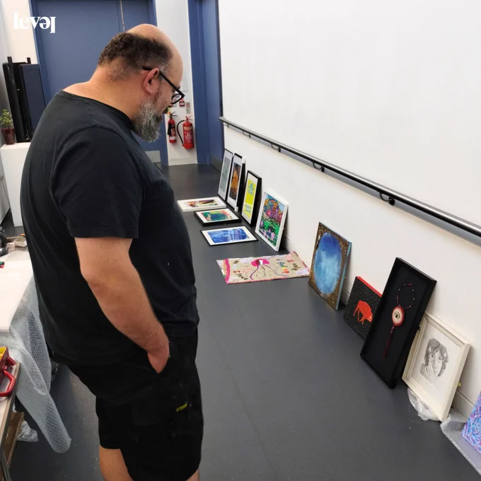 A person stands in a corridor looking down at some artwork that they are curating. The walls of the corridor are white and the floor is grey. There is a blue door at the end of the corridor.