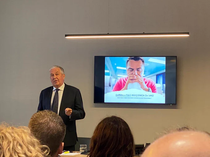 Speaker talking to a room of people in front of a screen showing a PowerPoint presentation
