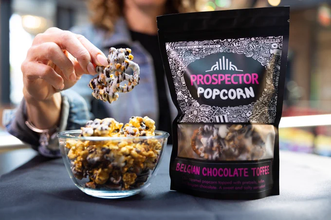 A woman holds up a chocolate covered pretzel that's clustered with toffee bits and caramel popcorn. A bowl of Belgian Chocolate Toffee gourmet popcorn sits next to a bag of the same flavor.