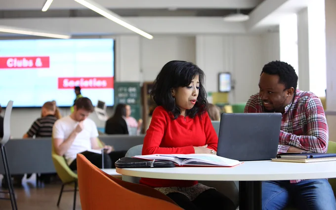 Two PhD students in conversation, sat at a table with laptop and notes