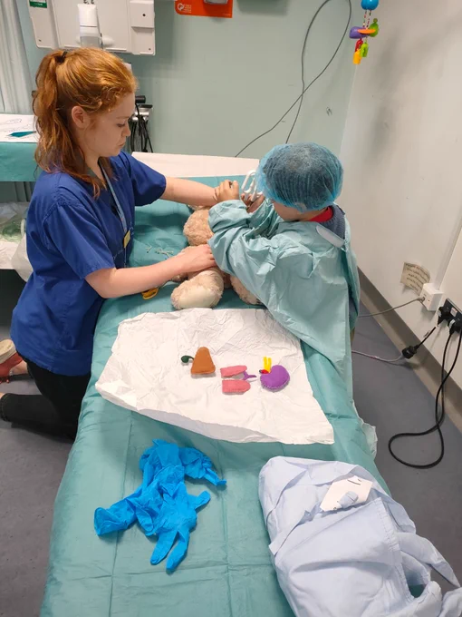 
A healthcare worker helps a child dressed in scrubs who is working on a teddy bear. There are organ shapes made of felt on the side.