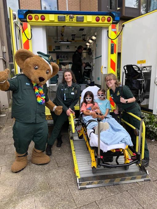 A person in a bear costume dressed in ambulance uniform stands with two other ambulance workers and two children at the back of an ambulance
