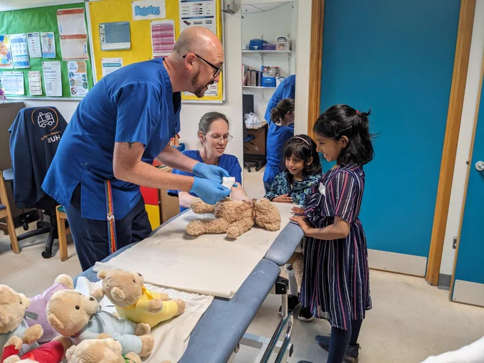 Two healthcare professionals apply a dressing to a teddy bear in front of two children