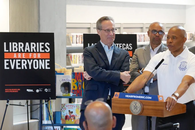 mayor stands at lectern inside library with signs next to him that read libraries are for everyone