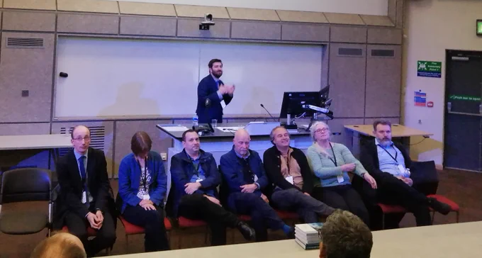 Seated panel of 5 men and 2 women. All smartly dressed and all nervous looking. Behind, Alex Nikitas is standing and enthusiastically introducing the panel