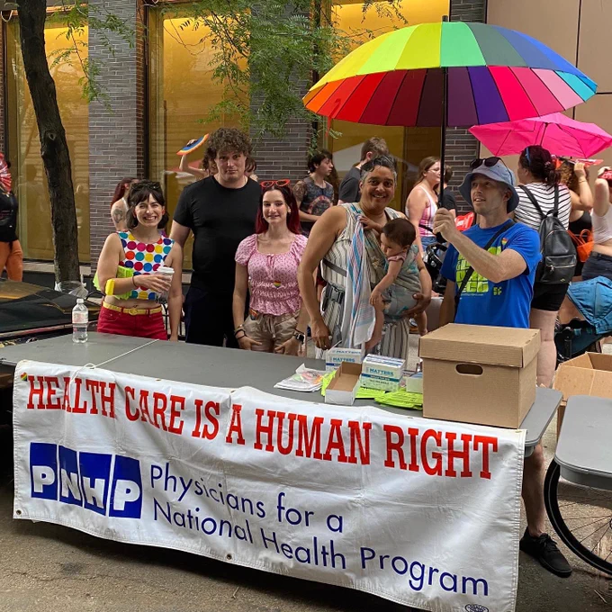 Part of our tabling team with rainbow outfits & umbrella at the Act Up health fair behind a folding table with the pnhp banner