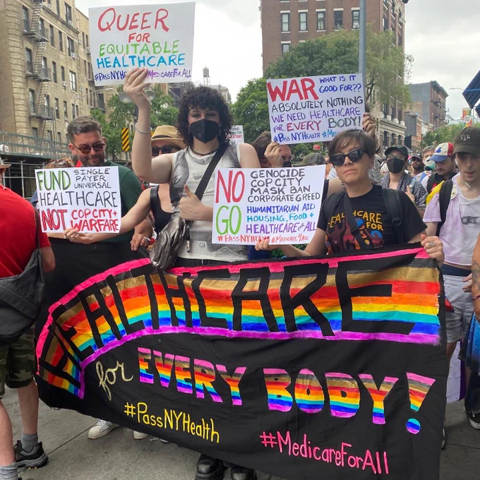 NY Health Act supporters holding a rainbow banner that says Healthcare for Every Body & signs calling for investment in universal healthcare not war