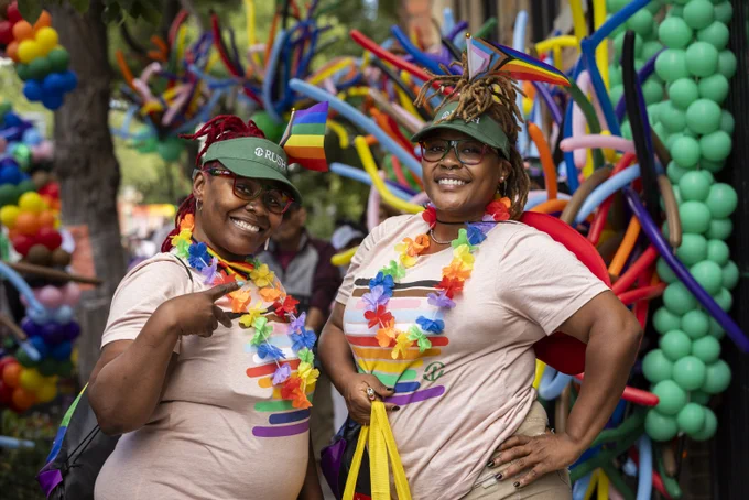 Two Rush staff members smiling at the Chicago Pride Parade, adorned with rainbow-themed accessories including hats, leis, and sunglasses, standing in front of a colorful background of multicolored balloons. One person gestures a peace sign with their hand.