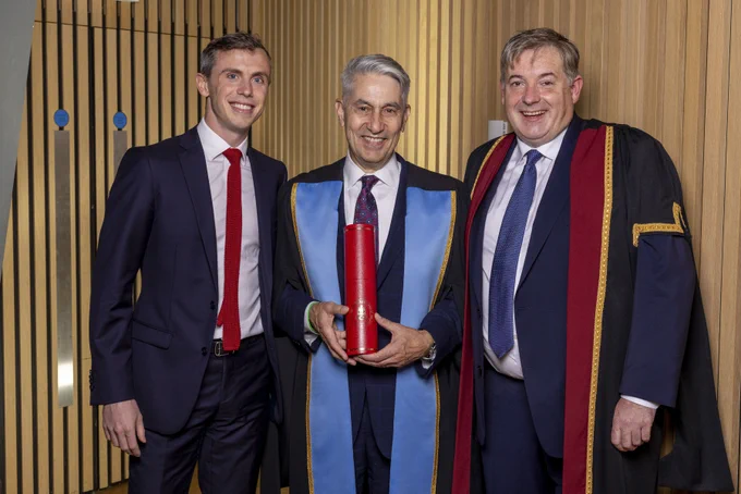 A group of three men stand. Two are wearing ceremonial robes over their suits. The man in the middle holds an RCSI scroll. In the background we see wood pannelling. 