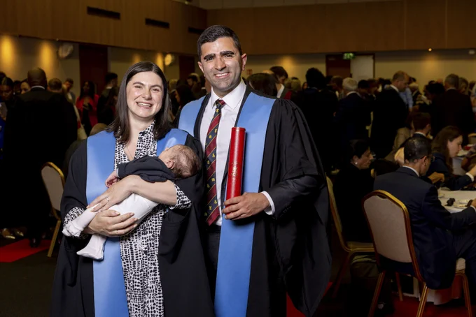 A couple stand in ceremonial robes. The woman on the left holds a tiny baby. The man on the right holds an RCSI scroll. Both are beaming. The baby is sound asleep.