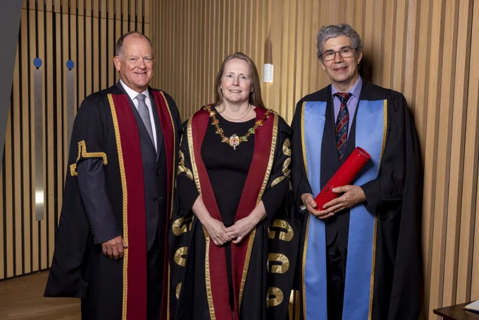A group of two men and a woman stand in front of wood panelling. All three are in ceremonial robes. The woman has a presidential chain. The man on the right holds an RCSI scroll.