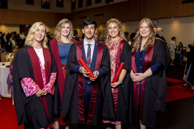 A group of five stand before a reception area. All are in ceremonial gowns of black and wine. Two hold RCSI scrolls. They are all beaming.
