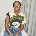 Photo of a woman in a tie-dye t-shirt sitting in a doctors office with a plaster on the top of her right arm.