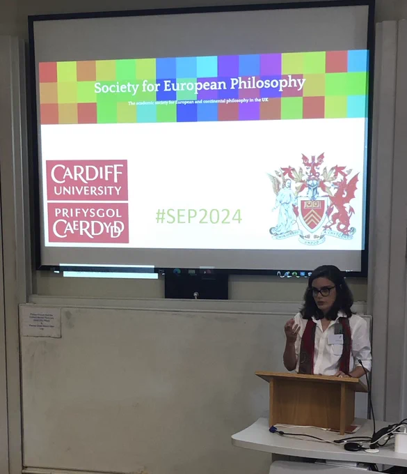 Juliana de Albuquerque speaking in front of a screen that presents the banner heading of the Society for European Philosophy and the logo and coat of arms of Cardiff University.