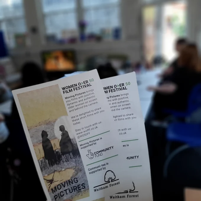 In the foreground of the picture, someone holds a flyer showing an illustration of 2 people talking at sunset. In the background of the picture, people sitting round a  table are watching a film
