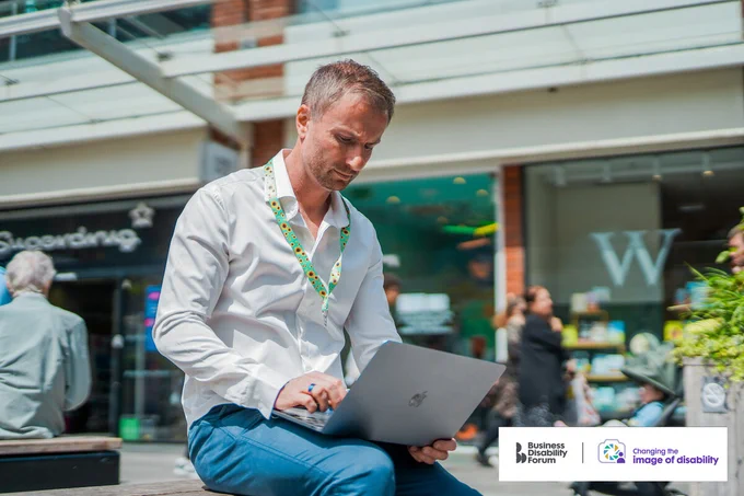 Person with a less-visible disability (early onset Parkinson's disease) wearing a sunflower lanyard uses a laptop at an outdoor shopping centre. The Business Disability Forum 'Changing the image of disability' logo is in the bottom right corner of the image.
