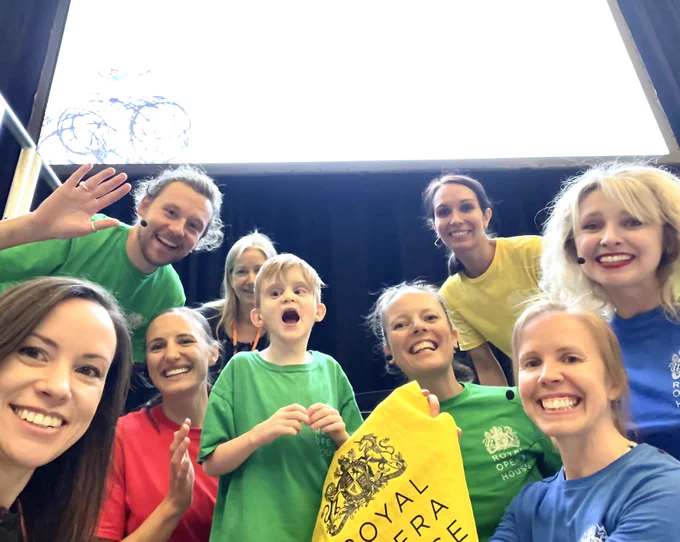 A selfie with 9 people smiling into the camera in front of a giant screen wearing brightly coloured T-shirts. The Royal Opera House logo can be seen in the front.