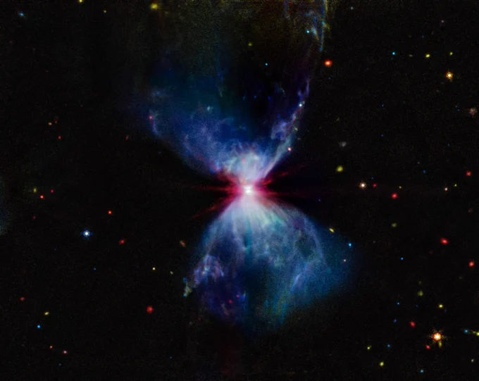 A growing protostar embedded within a molecular cloud. The center of the image shows a bright, red region, where the protostar resides, with a thin, gray lane cutting through it horizontally, which is the protostar’s accretion disk. Above and below this region are white and blue triangular-shaped cavities in the molecular cloud, which give the overall object an hourglass shape. The areas of the cavities closest to the central protostar have more pronounced plumes of white gas and dust that fade to a blue color further from the center. There are red, yellow, orange, blue, and green stars and galaxies scattered across the image.
