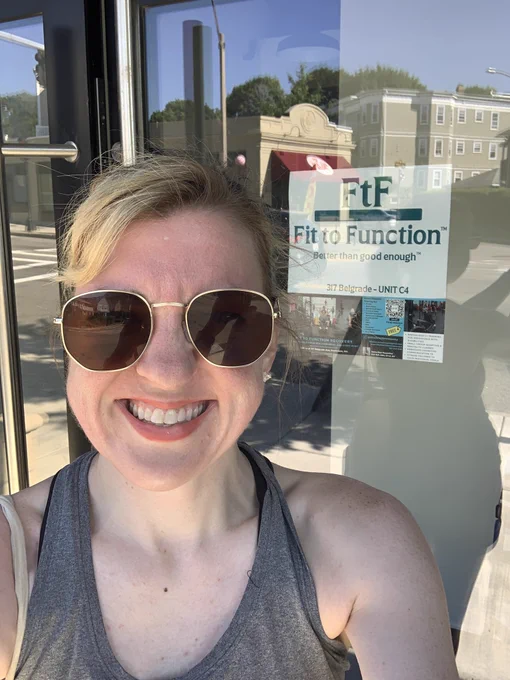 Blonde woman with aviator sunglasses and a gray tank top smiling outside of a gym sign that says Fit to Function. Better Than Good Enough