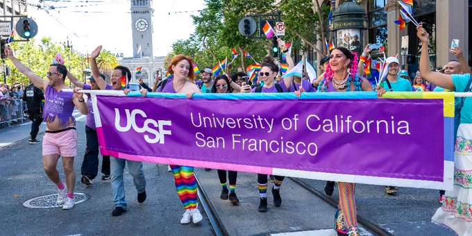 Group of people joyfully marching in a pride parade, holding a prominent 'University of California San Francisco' banner. They are surrounded by rainbow flags and a festive crowd.