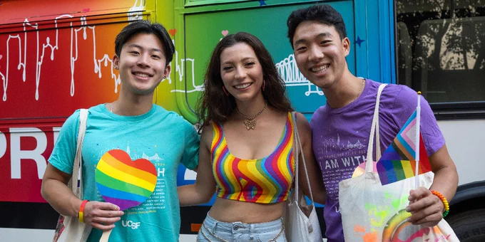 Three people smiling, standing in front of a colorful bus with a heart and rainbow design. They are wearing vibrant clothing, including T-shirts with rainbow motifs and a multicolored crop top. Each person is holding a small rainbow flag. The bus in the background displays the text "UCSF" and features blood drop designs and rainbow colors.