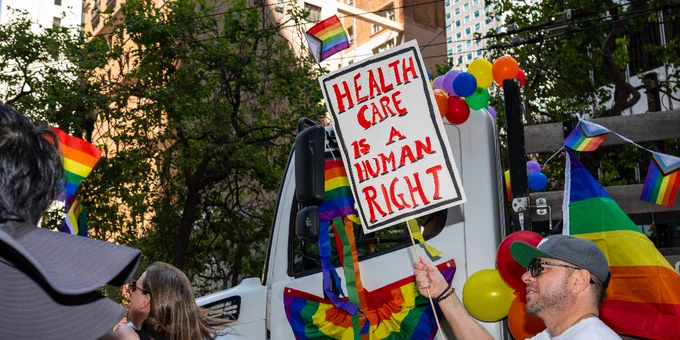 A person in sunglasses and a cap holds up a sign reading "HEALTH CARE IS A HUMAN RIGHT" at a Pride event. The sign is colorful, decorated with rainbow stripes, and surrounded by balloons and flags also featuring rainbow colors.