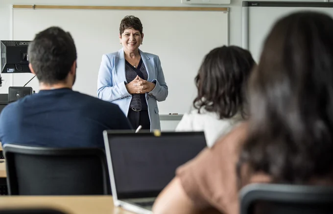 A woman in a blue blazer stands teaching a classroom of college students taking notes on their laptops.