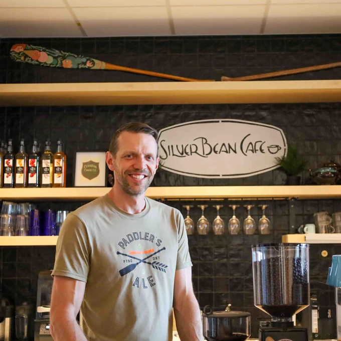 Dan Brandsma, the Silver Bean Cafe owner, stands in front of the new cafe location at the museum and smiles. Behind are painted paddles, the Silver Bean sign, and cafe dishware.
