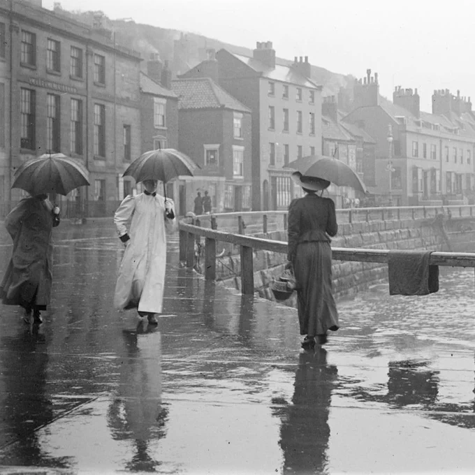 A black and white photograph of people walking in the rain on the seafront with umbrellas. 