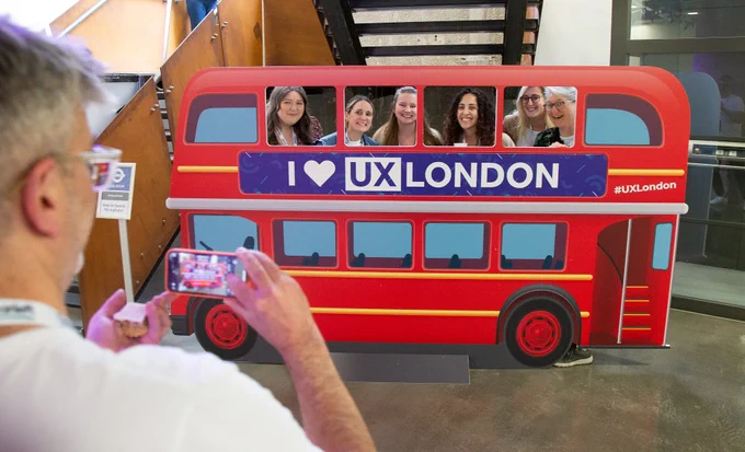 A man photographs a group of five women smiling and posing behind a cutout of a double-decker bus at an event. The cutout has the text "I ♥ UX London" on it. The scene takes place indoors, likely at a conference or similar gathering.