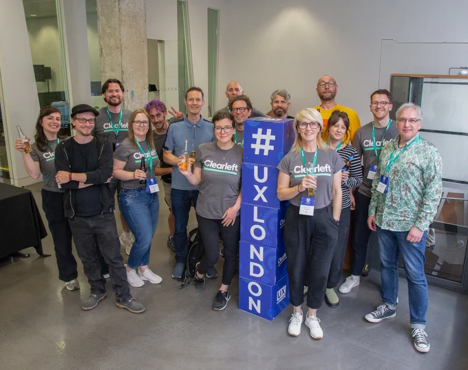 A group of people posing and smiling for a photo around a blue #UXLONDON sign. Some are holding drinks, and many are wearing conference badges and Clearleft t-shirts. The background shows a modern interior with glass walls and concrete pillars.