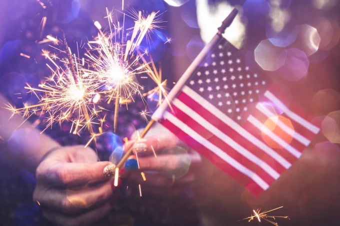 Person holding a sparkler in one hand and a small American flag in the other, with a backdrop of blurred lights and bokeh effect.