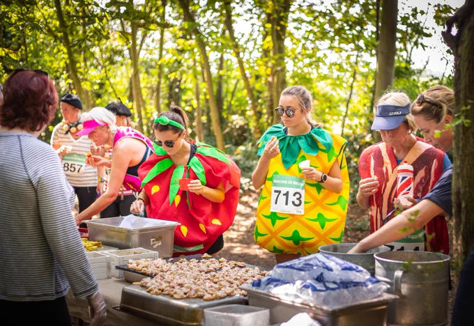 Runners stopping at the food stop in the woods on the 10k course, dressed as pineapples, bacon and a strawberry for the Marathon du Malton 10k run. 