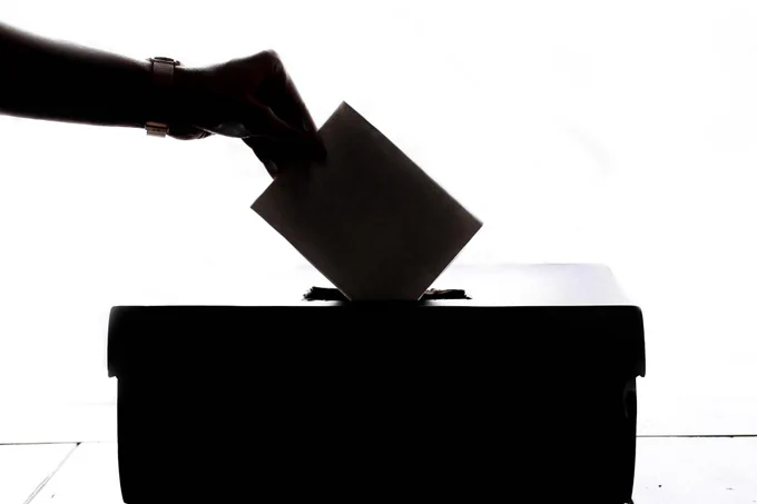 A silhouette of a hand putting a folded up  piece of paper into a box traditionally used for voting.