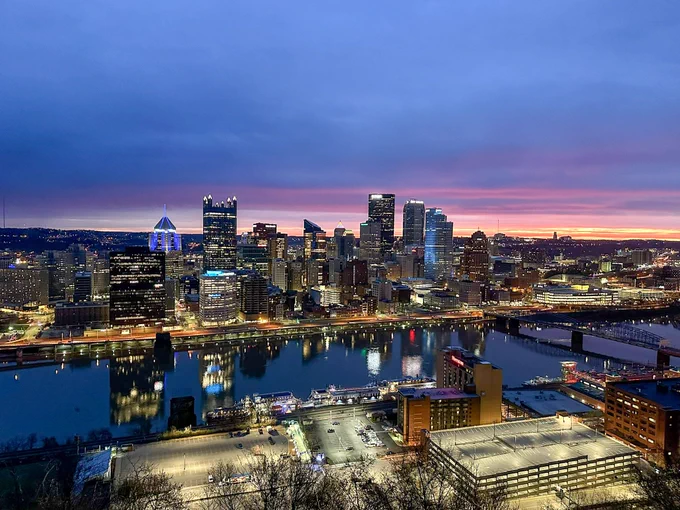 Aerial view of a cityscape at twilight featuring illuminated high-rise buildings and a river, with a vibrant sunrise in the background.