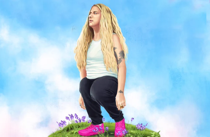 Short woman with long blonde hair standing on small patch of grass with purple flowers. Blue sky in background. 