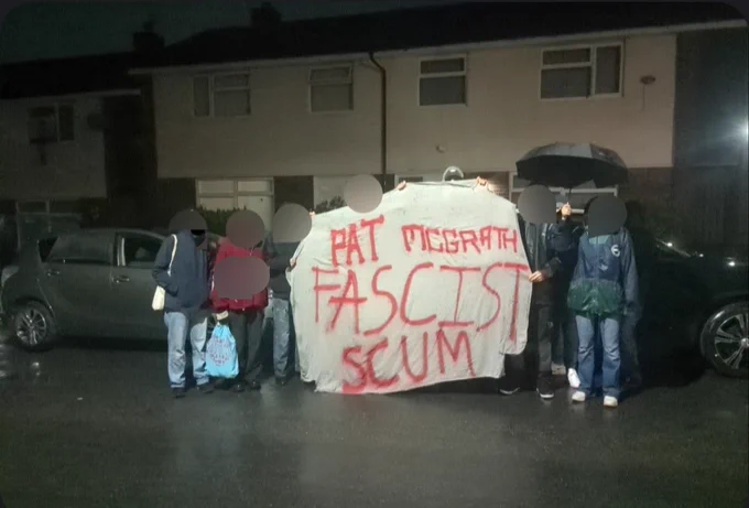 A crowd of antifascists outside Patrick McGrath's house with a banner that reads 'PAT MCGRATH FASCIST SCUM' in red paint.
