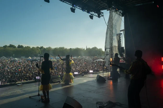 A band performs on a large festival stage in a park setting whilst a large festival audience watches. The sky is blue and there's large green trees all around.