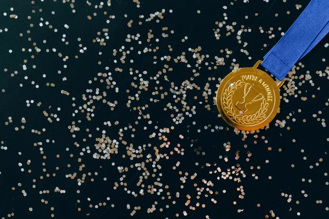A gold medal with a blue ribbon lies on a dark surface scattered with small, round, gold and silver confetti. The medal is engraved with the phrase "You're A Winner" and has an image of a trophy surrounded by laurel leaves.