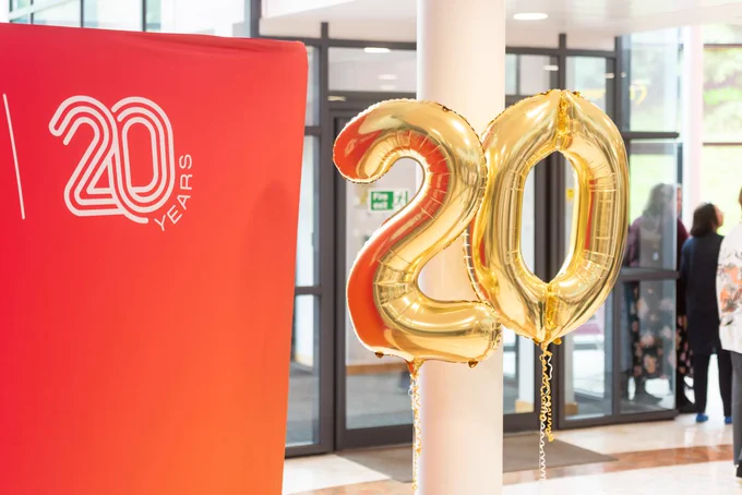 Banner displaying "20 years" beside golden balloons shaped as the number "20" in a brightly lit indoor setting with people in the background.