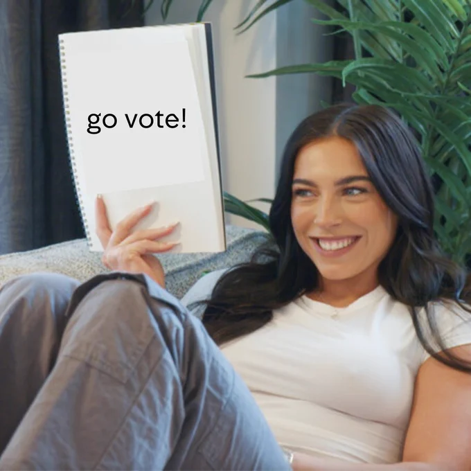 An image of Erica from Married At First Sight UK. She has dark brown hair and is wearing a white T-shirt and grey cargo trousers. She's sitting on a sofa smiling holding up a sign that says 'go vote!"