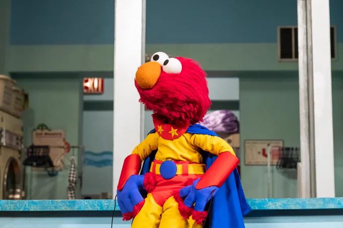 Elmo is a super hero costume, standing with his hands on his hips looking to the side majestically