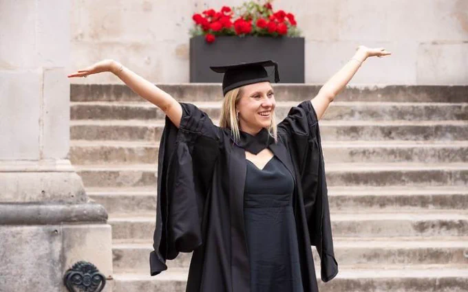 A graduate happily celebrating on stairs, arms spread wide, wearing a black graduation gown and mortarboard. T