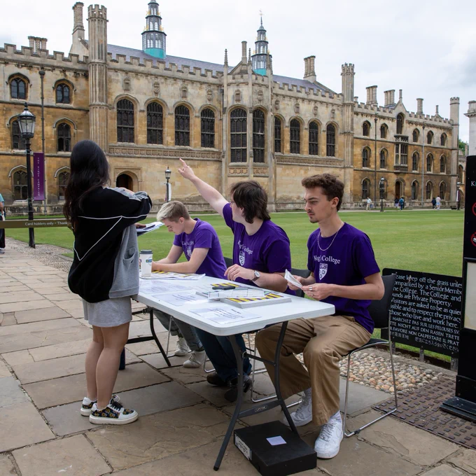 Three individuals in purple shirts manning a desk in the historical grounds of King's College, Cambridge. 