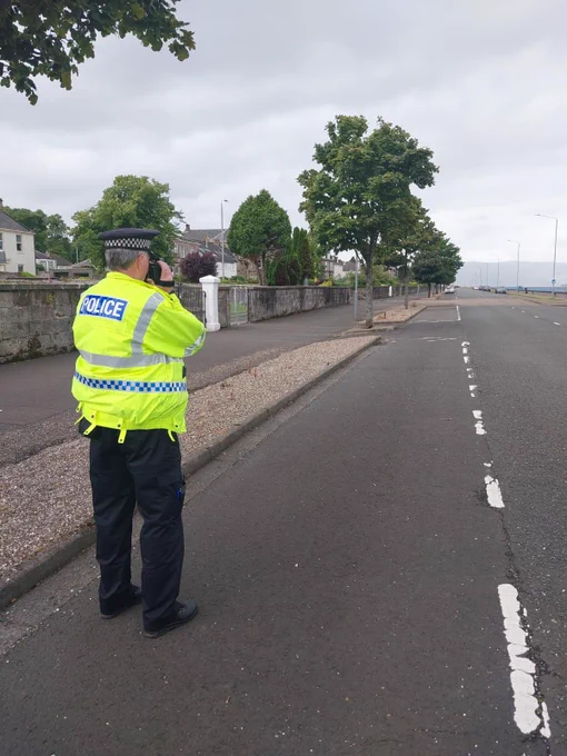 Police Officer standing on roadway with back to the camera, yellow flourescent jackets on, holding speed checking device pointing down the road.