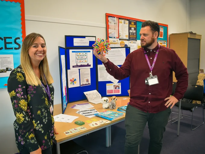 Two people stand by a display board covered in educational materials and posters, in a classroom setting. One person is holding a colorful, spherical model, while both appear engaged in an enthusiastic discussion.